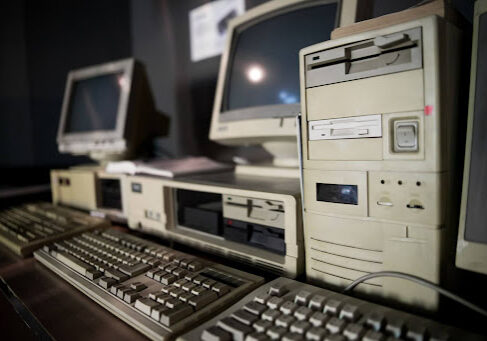 A row of vintage desktop computers with CRT monitors, keyboards, and floppy disk drives on a desk.