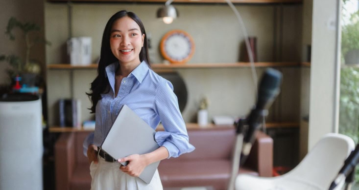 Smiling young Asian woman holding laptop in modern home office.